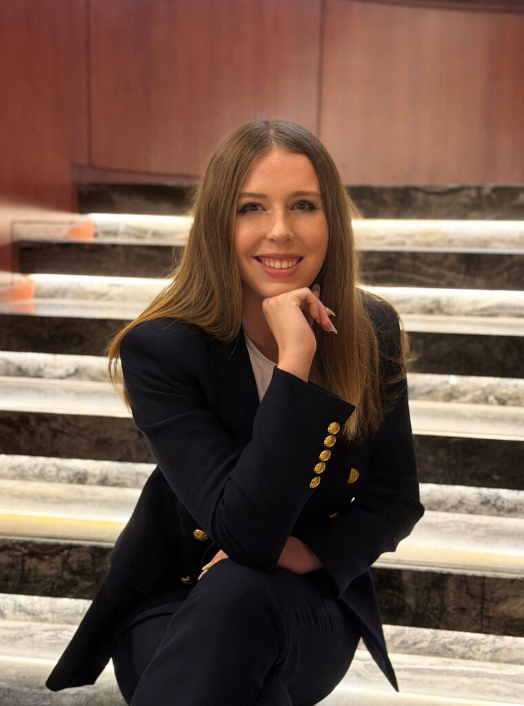 Grace Dunn seated on marble stairs in a navy blazer with a warm smile — National Merit Scholar and elite PSAT tutor at Crownridge Coaching